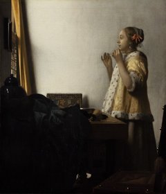 Eine junge Frau steht an einem Tisch und blickt in einen Spiegel, um sich eine Kette anzulegen