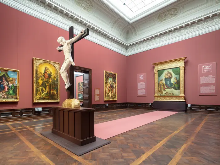 Eine Ausstellungsansicht mit einem Kruzifix, der vor der "Sixtinischen Madonna" steht. 